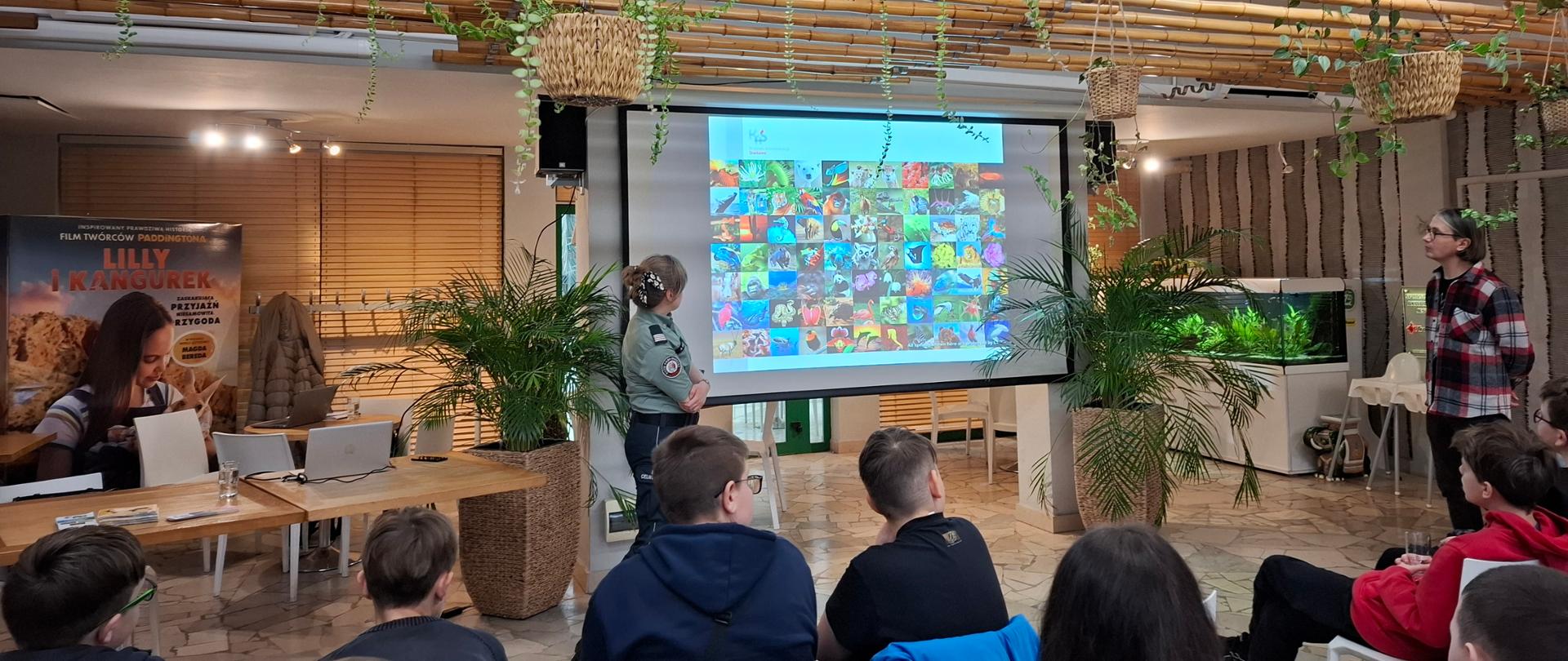 Grupa dzieci w wieku ok. 13 lat patrzy z zainteresowaniem na ekran - prelegentka w mundurze służby celno-skarbowej opowiada o zagrożonych zwierzętach.
