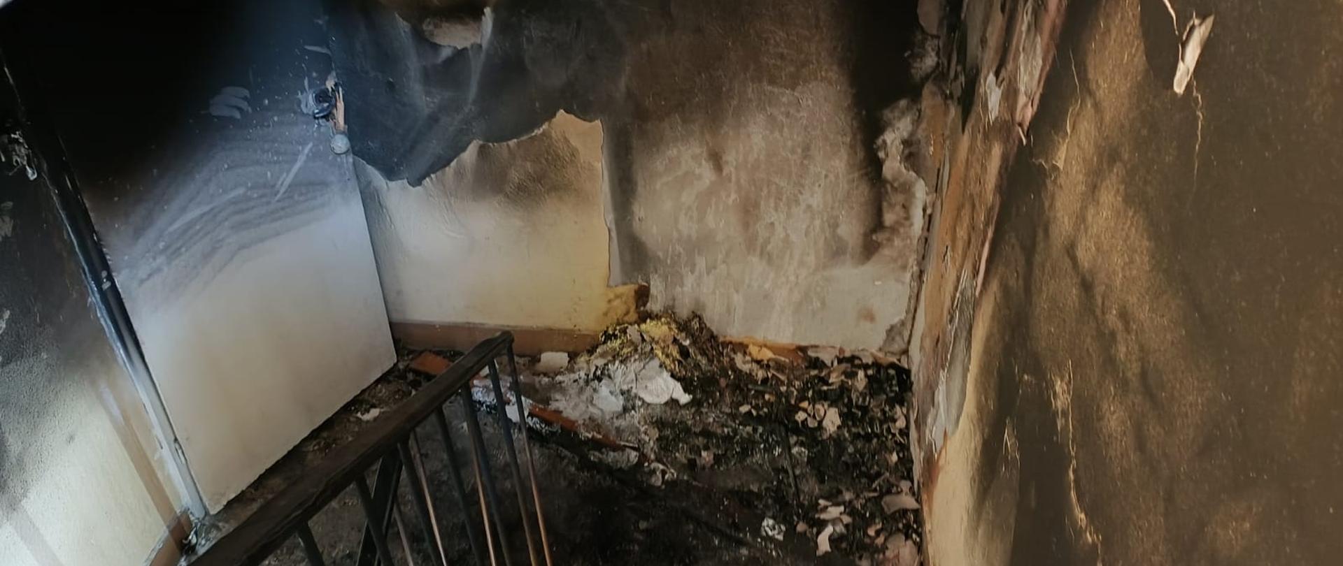 Zdjęcie przedstawia zniszczoną przez ogień klatkę schodową. Okopcone są tynki sufitu i ścian, widać też ubytki tynku spowodowane działaniem temperatury.