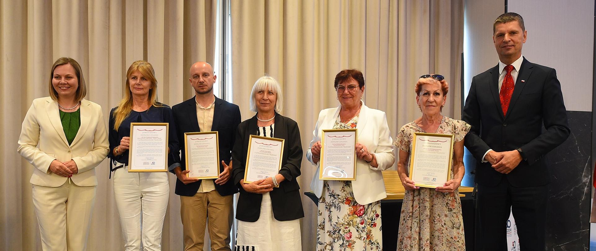 Wiceminister Piontkowski stoi w szeregu ludzi trzymających oprawione w ramki dyplomy.