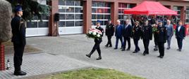 Składanie kwiatów pod obeliskiem pamiątkowym na placu komendy. Po lewej stronie na baczność stoi strażak w mundurze galowym, pełniąc wartę przy kamiennym obelisku. Środkiem placu maszeruje strażak niosący duży wieniec z białych kwiatów z biało-czerwoną wstęgą. Za nim w kolumnie idą zaproszeni goście, w tym komendanci w mundurach oraz przedstawiciele władz w garniturach. W tle widoczny budynek straży pożarnej i czerwony namiot.