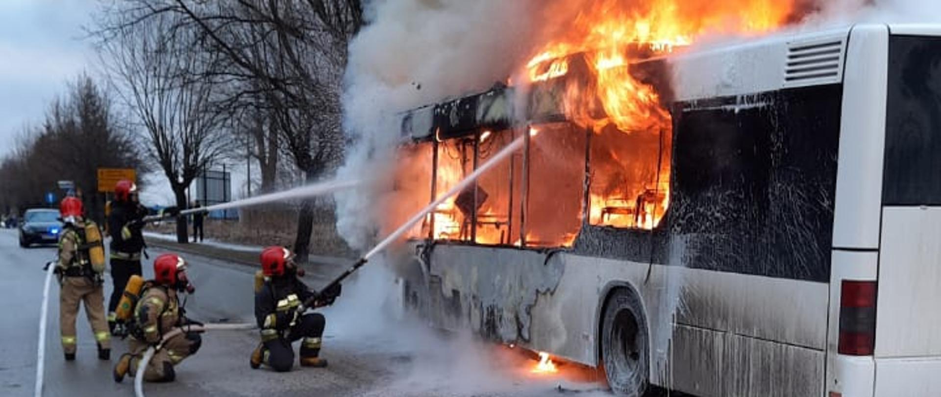 Na zdjęciu widać palący się kursowy autobus komunikacji miejskiej. Płomienie wydostają się na zewnątrz pojazdu przez okna. Strażacy ubrani w ubrania specjalne i aparaty ochrony układu oddechowego podają dwa prądy piany gaśniczej na źródło pożaru. Pożar obejmuje niemalże cały pojazd. Kłęby dymu wydostające się z pożaru przysłaniają rosnące z tyłu drzewa.