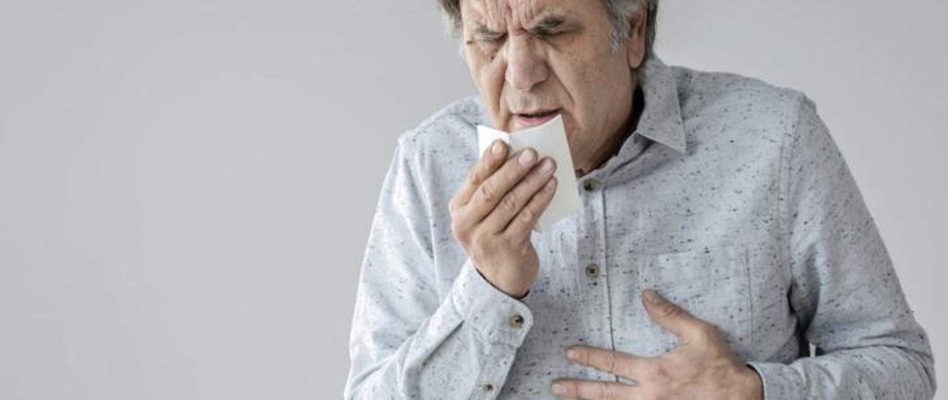 Starszy mężczyzna kaszle w chusteczkę a drugą ręką trzyma się za piersi 
