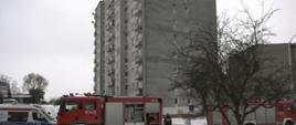 Na pierwszym planie samochody strażackie i karetka pogotowia, w tle budynek mieszkalny wielorodzinny w którym doszło do pożaru. 