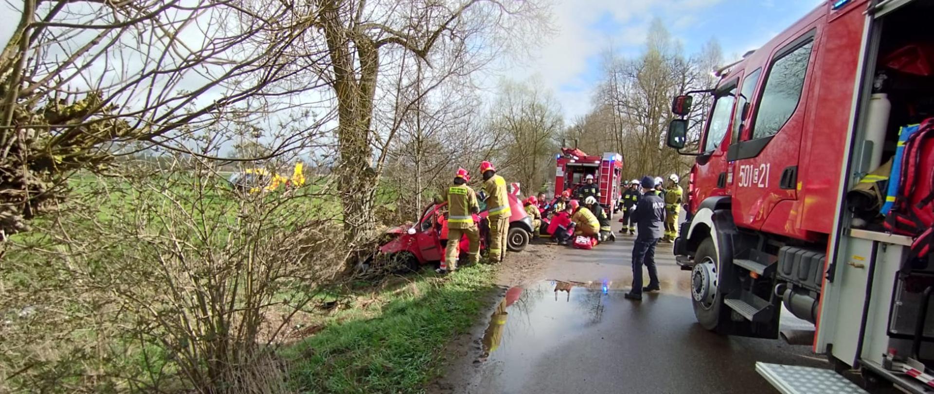 Dwa wozy strażacki stoją na drodze, samochód osobowy z rozbitym przodem uderzył w drzewo, przy wypadku stoją strażacy, ratownicy medyczni i policjanci . na polu stoi żółty helikopter pogotowia
