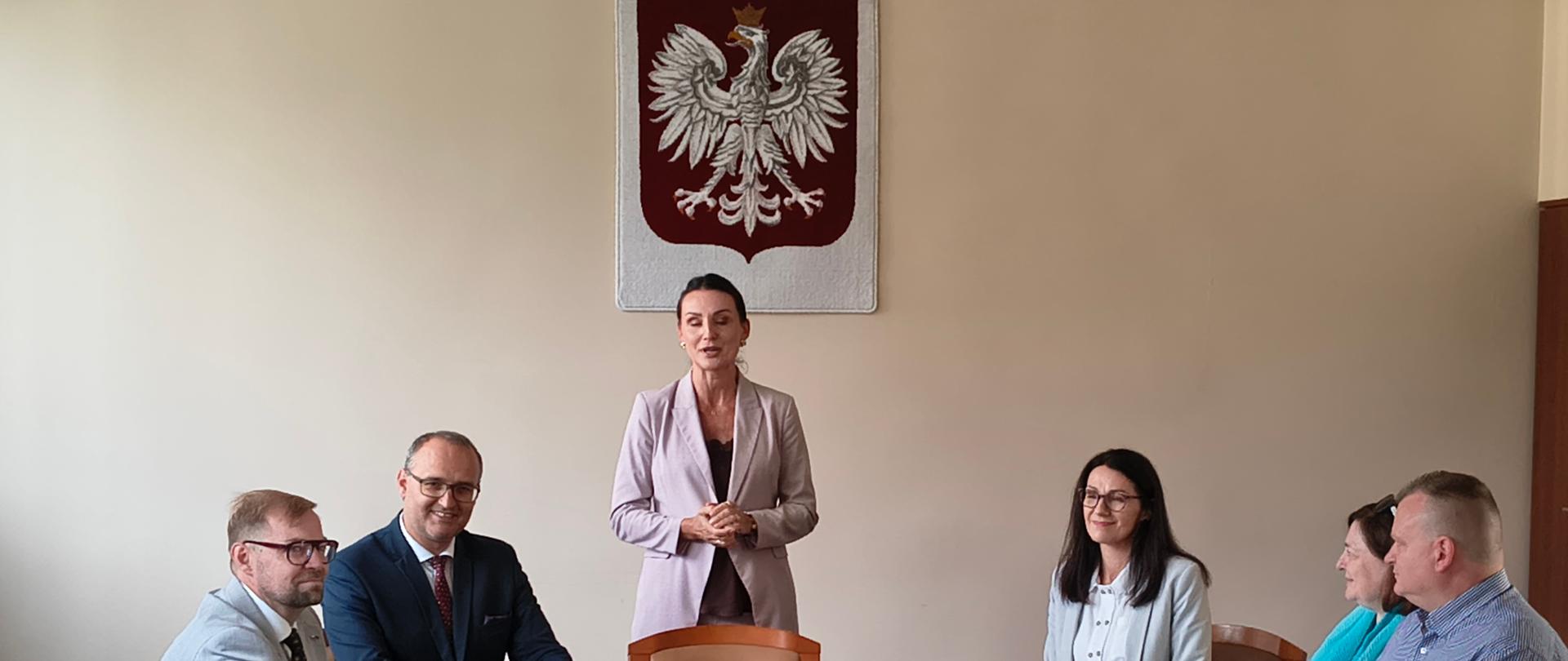 Grupa osób siedzi przy owalnym stole patrząc na kobietę, która stoi przy stole na tle godła polskiego 