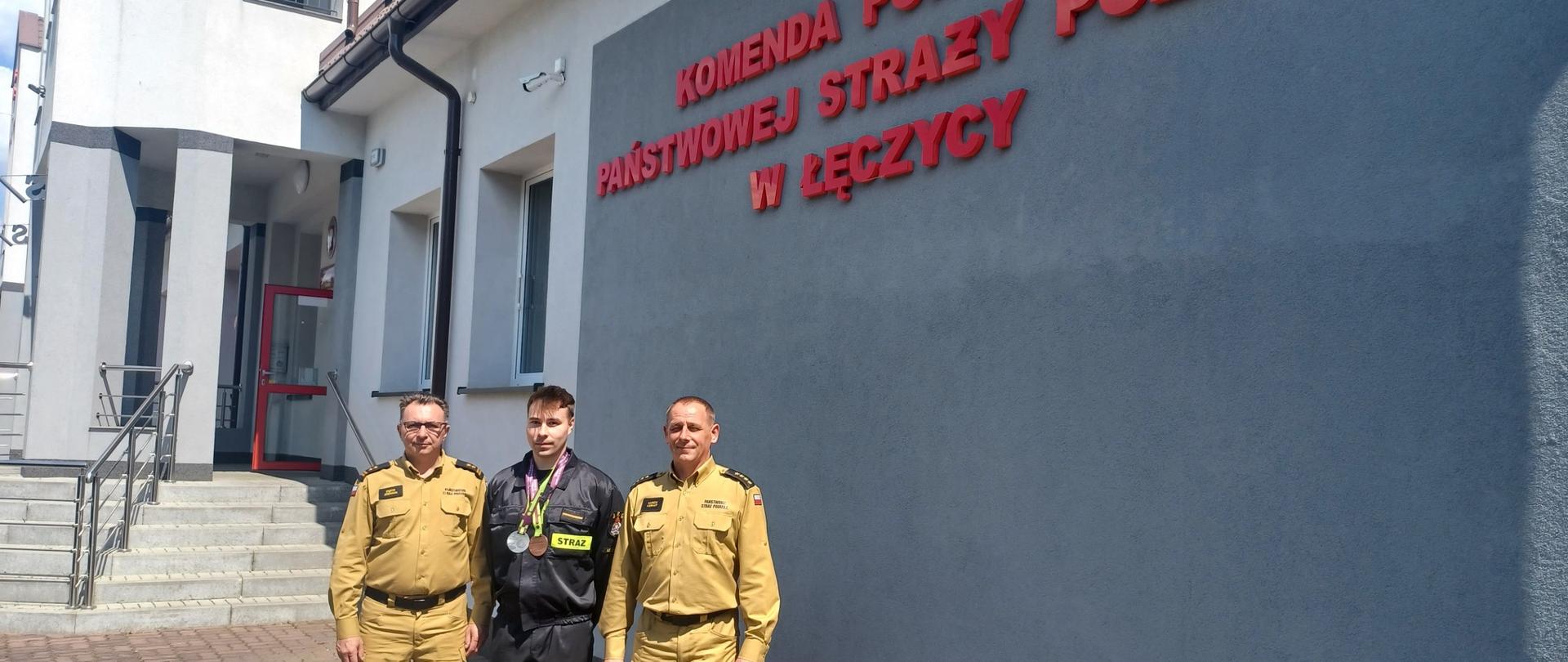 Na zdjęciu widać trzech strażaków, którzy pozują do zdjęcia. Dwóch ubranych jest w żółte stroje. Po środku stoi trzeci w czarnym stroju. Na szyi ma zawieszone medale. W tle widać budynek. Na nim jest napis Komenda Powiatowa państwowej Straży Pożarnej w Łęczycy . Jest widno.