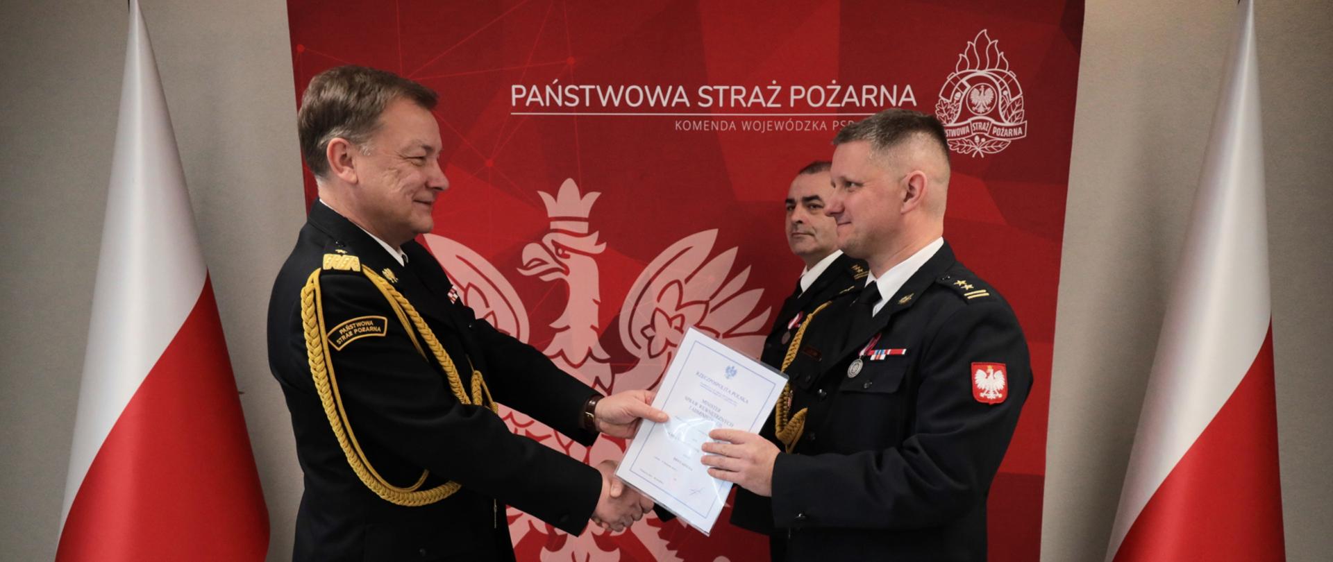 Strażak w mundurze wyjściowym wręcza dokument bryg. Łukaszowi Olejnikowi podczas uroczystości. Obaj stoją na tle czerwonej ścianki z godłem Polski i logo Państwowej Straży Pożarnej. W tle widoczny jest Komendant Powiatowy PSP w Kluczborku. Wszyscy w umundurowaniu wyjściowym. Po bokach ustawione są flagi Polski.