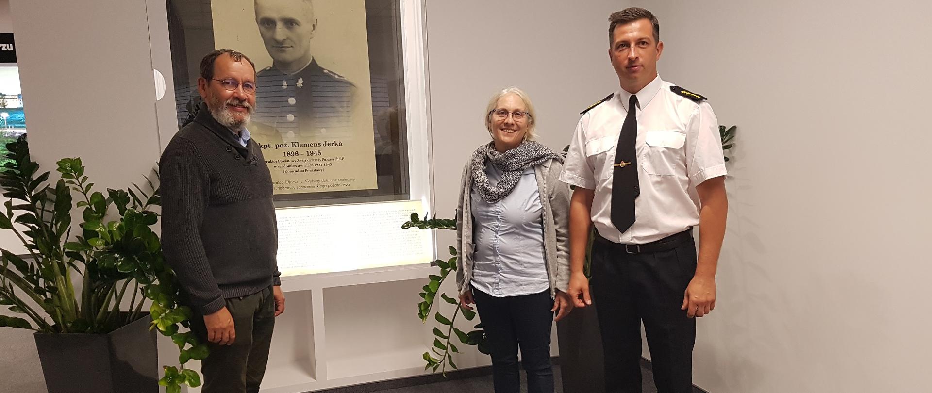 Zdjęcie zrobione w reprezentacyjnej Sali Komendy. Przedstawia trzy osoby stojące przy miejscu pamięci o kpt. Jerce. Jeden z mężczyzn stoi po lewej stronie a kobieta i strażak w mundurze stoją po prawej stronie. W środku znajduje gablotka ze zdjęciem i notą biograficzna kapitana Jerki.