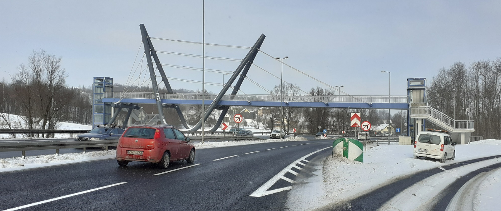 nad drogą widać kładkę dla pieszych, pod kładką przejeżdża samochód, po lewej zjazd w prawo