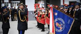 Nadbryg. Grzegorz Szyszko oraz inni oficerowie PSP salutują pocztowi sztandarowemu. W tle widoczna orkiestra w czerwonych mundurach, co podkreśla uroczysty charakter wydarzenia.