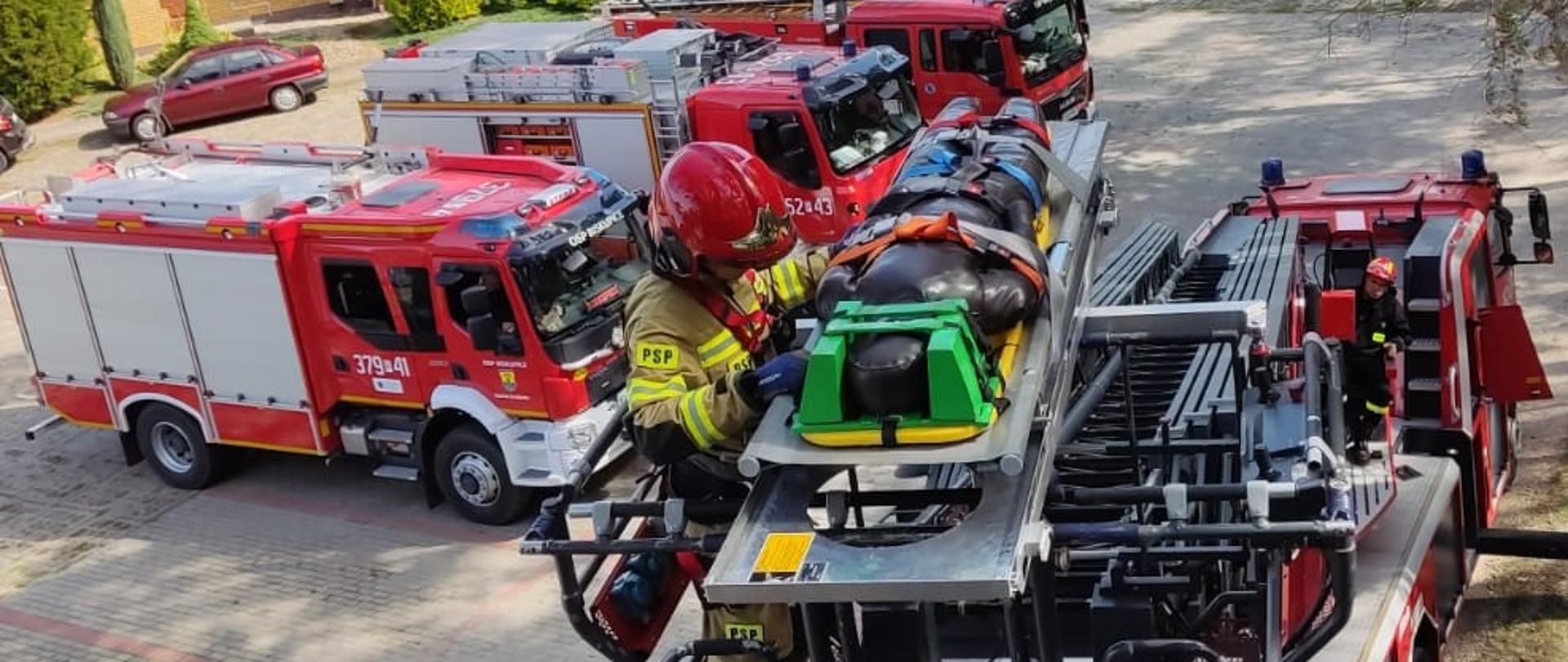 Strażak w koszu drabiny mechanicznej, na wysokości, asekuruje poszkodowanego (manekin) leżącego na noszach. W tle widać 3 wozy strażackie oraz samochód cywilny, drzewa i budynek. 