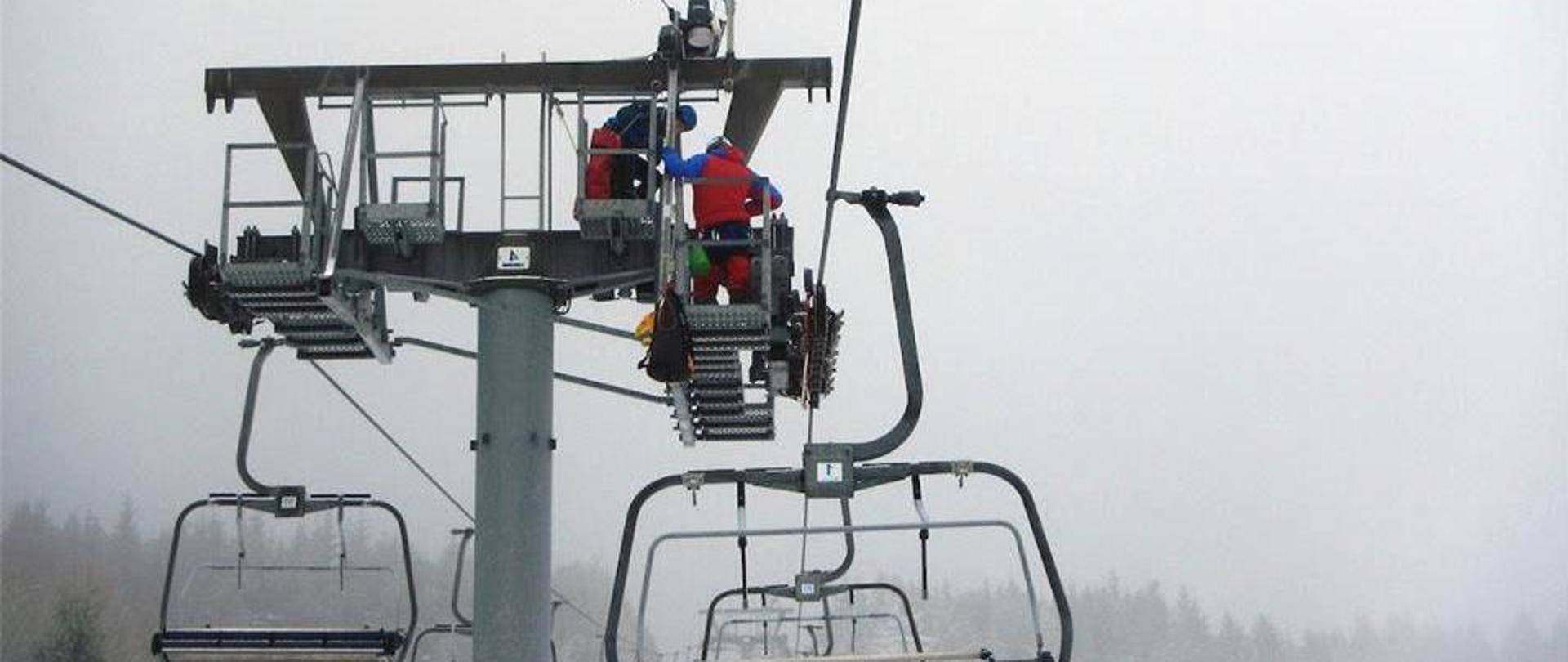 Zdjęcie przedstawia krzesełkowy wyciąg narciarski na którym strażacy Państwowej Straży Pożarnej ubrani w czerwone kombinezony do ratownictwa wysokościowego prowadzą ćwiczenia z ewakuacji. Dwóch ratowników znajduje się na szczycie słupa podtrzymującego liny wyciągu