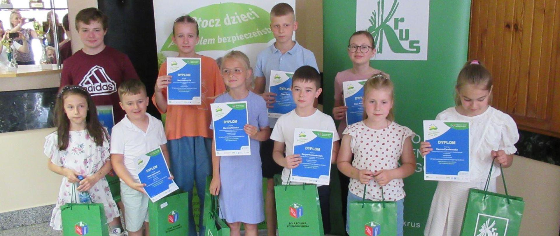Grupa dzieci z dyplomami na tle rollupów z logo KRUS.