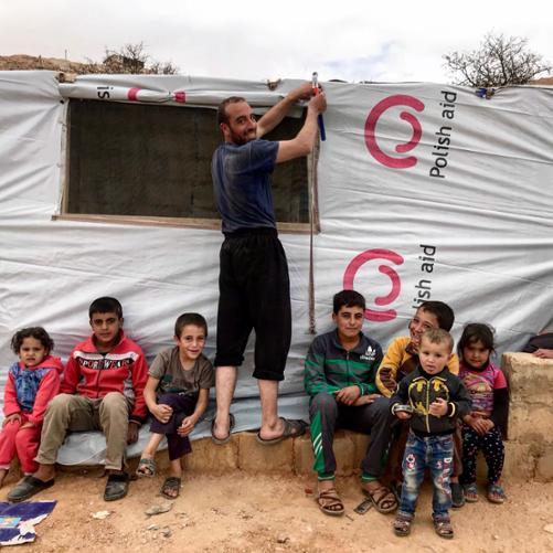 mężczyzna zakłada płachtę na namiot, a u jego stóp siedzą dzieci