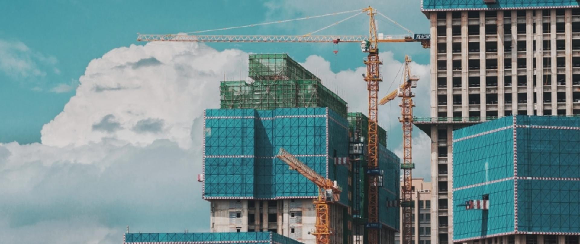 Budowa z dźwigami i budynkami w trakcie realizacji, osłoniętymi niebieskimi siatkami.