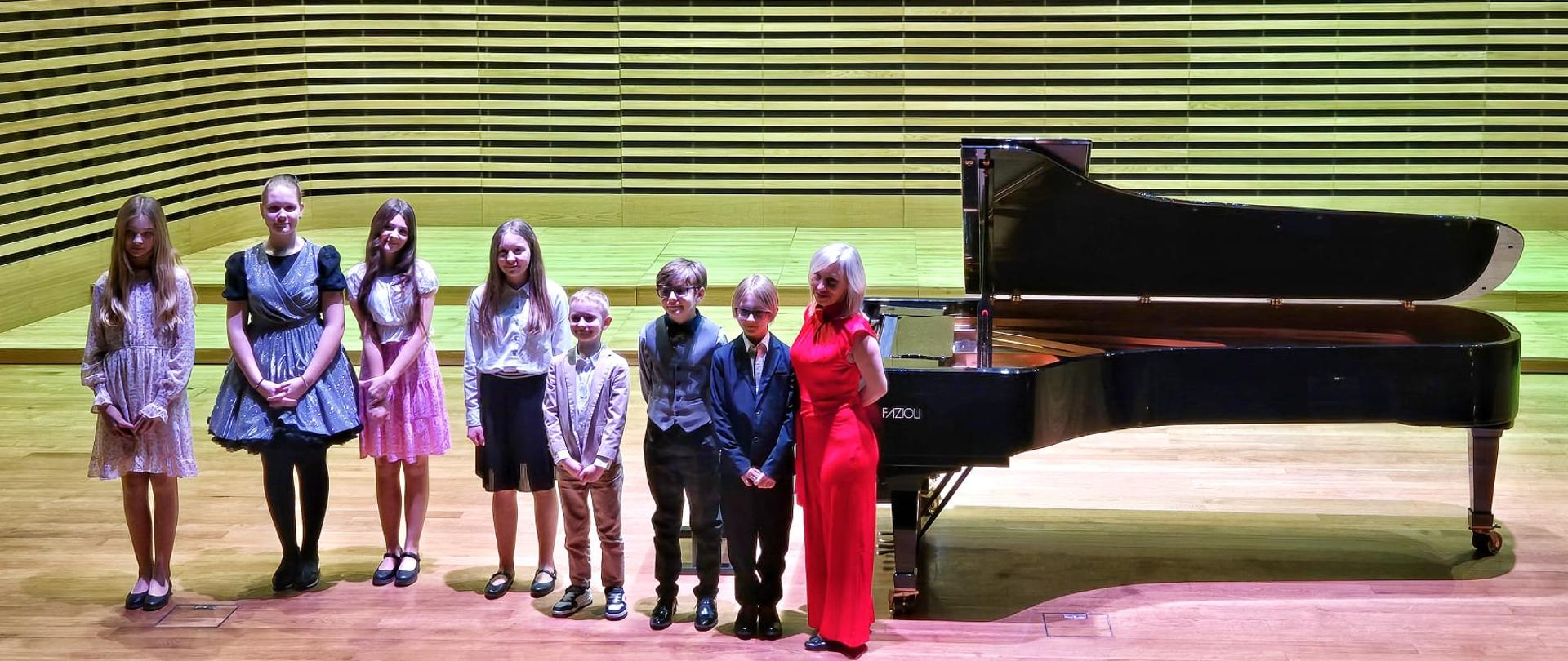Nauczycielka z uczniami swojej klasy stoją na scenie obok fortepianu.