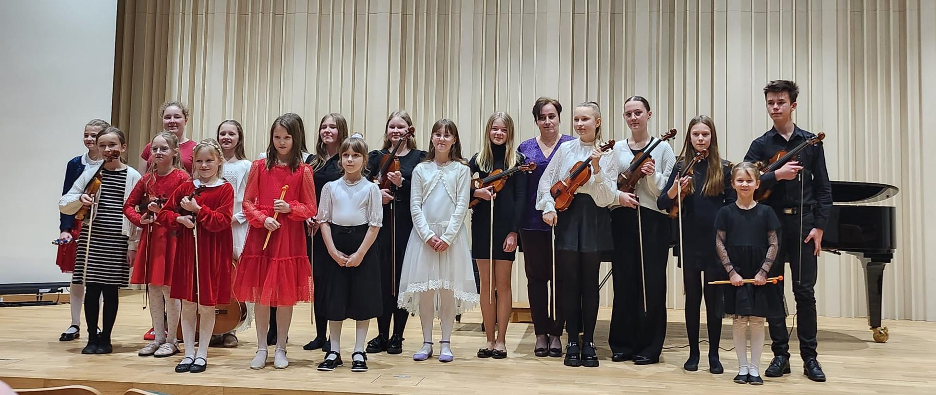 Na zdjęciu grupa odświętnie ubranych uczniów. W tle za nimi czarny fortepian i panele w odcieniach brązu. Pamiątkowe zdjęcie młodych artystów, którzy wystąpili podczas rodzinnego kolędowania dla babci i dziadka.