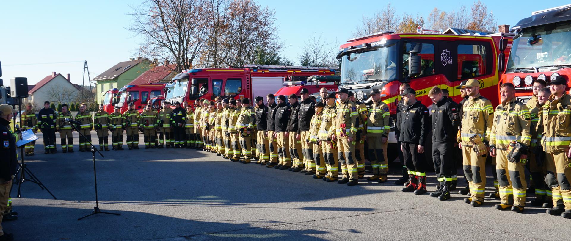 Zdjęcie przedstawia około 50 strażaków ochotniczej straży pożarnej ubranych w ubrania bojowe, którzy stoją przed samochodami strażackimi. Jest to odprawa przed ćwiczeniami, strażacy stoją w dwuszeregu są zwróceni w jedną stronę.