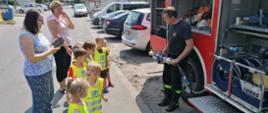 Zdjęcie przedstawia dzieci z Przedszkola Akademia Wiedzy i Zabawy wraz z opiekunami przy samochodzie strażackim. Strażak omawia i prezentuje sprzęt pożarniczy.