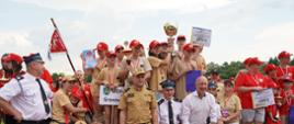 Grupka dzieci w koszulach w kolorze piaskowym stoi na podium. Dzieci na szyjach mają założone medale, a kilka z nich trzyma duży puchar. Przy podium stoi kilka osbó dorosłych, w tym funkcjonariusz PSP w piaskowym ubraniu służbowym i czapce z daszkiem, kilku druhów OSP ora jedna osoba cywilna.