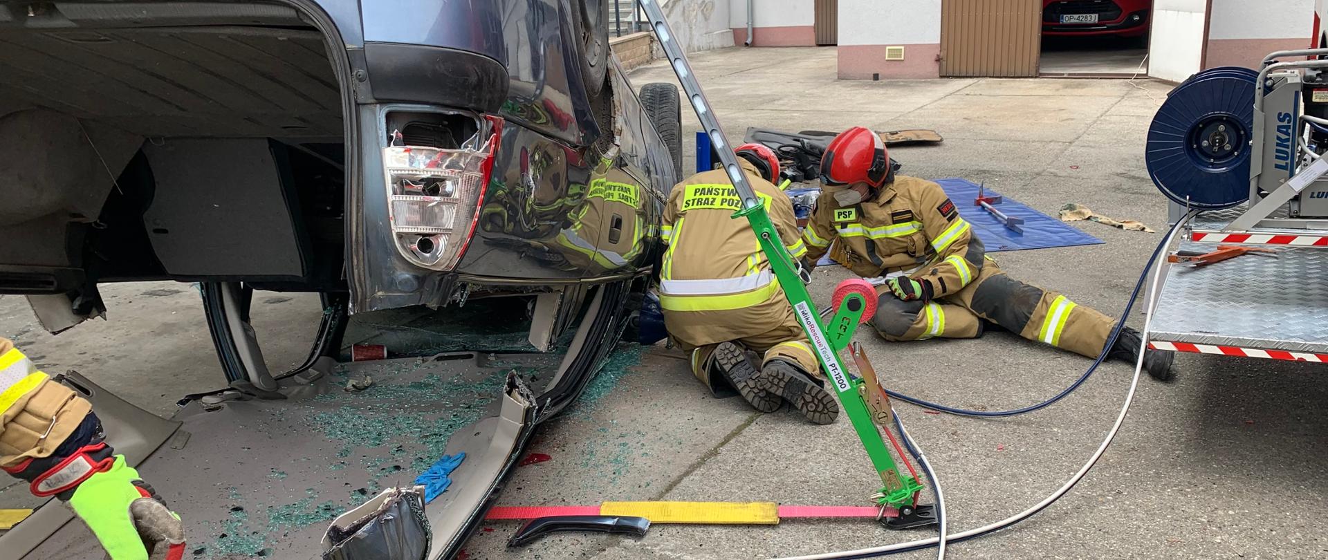 Po lewej samochód osobowy leżący na dachu, uwidoczniony od tyłu. W samochodzie częściowo odcięto dach, który leży teraz pod nim. Samochód ustabilizowano podporami. Po prawej stronie dwaj strażacy klęcząc odcinają przedni słupek pojazdu. Ratownicy w ubraniach specjalnych w kolorze piaskowym i czerwonych hełmach. 