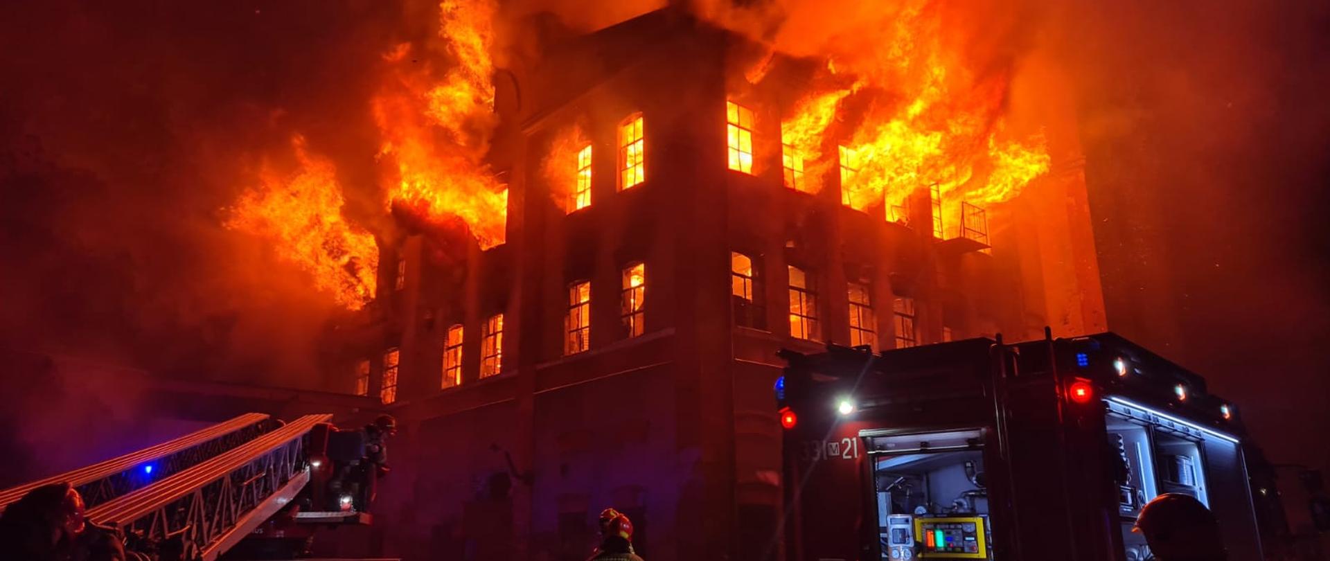 Zdjęcie wykonane w porze nocnej. Zdjęcie przedstawia pożar nie użytkowanej hali produkcyjno – magazynowej w Radomiu przy ulicy Młodzianowskiej. Budynek trzy piętrowy. Na pierwszym i drugim piętrze z powybijanych okien wydobywają się na zewnątrz dym i płomienie. Przed budynkiem znajdują się pojazdy ratowniczo – gaśnicze: od lewej pojazd drabina mechaniczna oraz dalej po prawej stronie pojazd gaśniczy. Pomiędzy samochodami znajdują się również strażacy uczestniczący w akcji gaśniczej. Zdjęcie wykonane w przybliżeniu w porównaniu do zdjęcia numer dwa. 