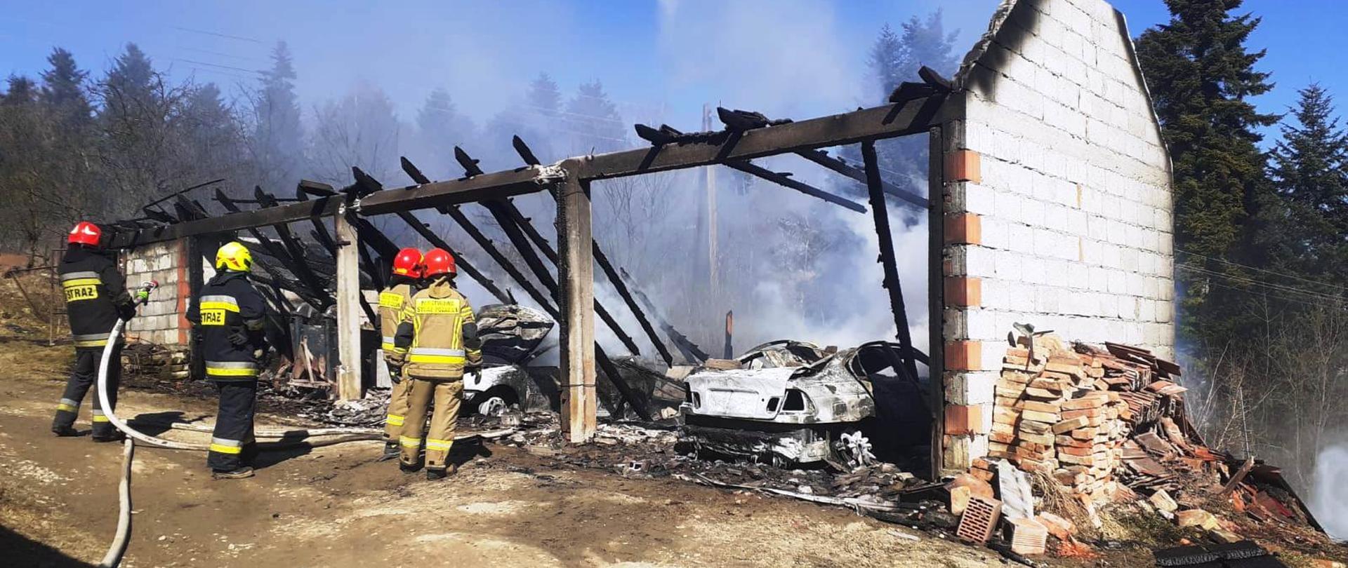 Strażacy gaszą pożar budynku gospodarczego.Wewnątrz widać spalone samochody.