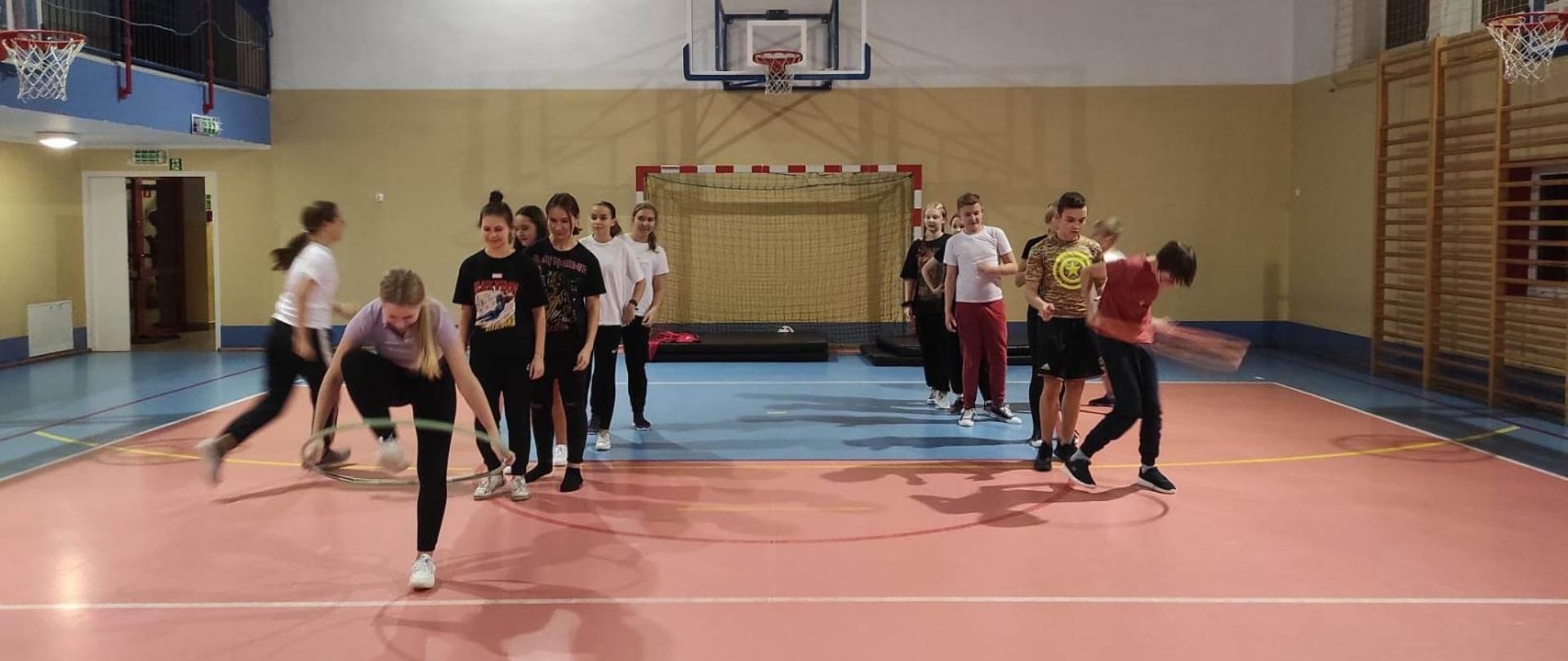 Na sali gimnastycznej stoją nastolatkowie w dwóch rzędach. Pierwsze osoby w rzędzie skaczą przez koło hula-hop.
