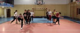 Na sali gimnastycznej stoją nastolatkowie w dwóch rzędach. Pierwsze osoby w rzędzie skaczą przez koło hula-hop.