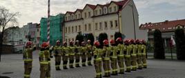Zdjęcie przedstawia strażaków stojących na placu przed budynkiem straży pożarnej podczas uroczystego wciągnięcia flagi na masz. 