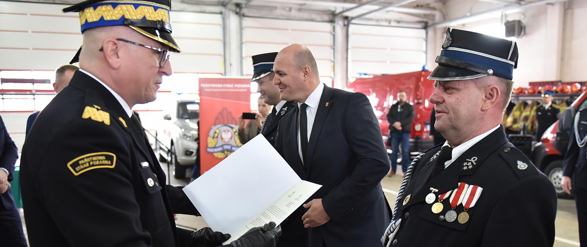 na głównym planie generał straży pożarnej wręcza decyzję strażakowi ochotnikowi, w tle inne osoby, zdjęcie wykonane w garażu