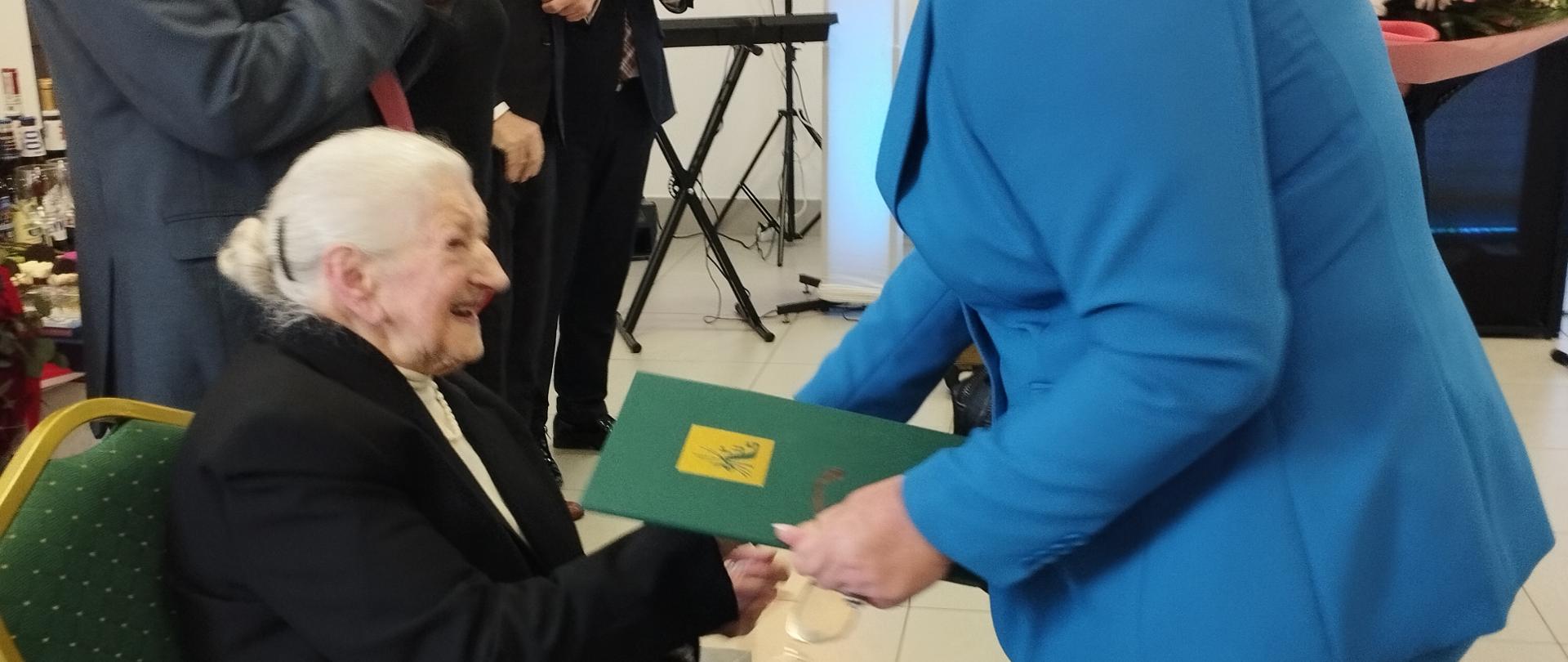 na krześle siedzi Jubilatka Pani Helena Kuchta. Obok stoją członkowie rodziny. Naprzeciwko stoi Kierownik Placówki Terenowej KRUS w Pułtusku Anna Łojewska i Kierownik Placówki Terenowej KRUS w Ciechanowie Jacek Biliński.