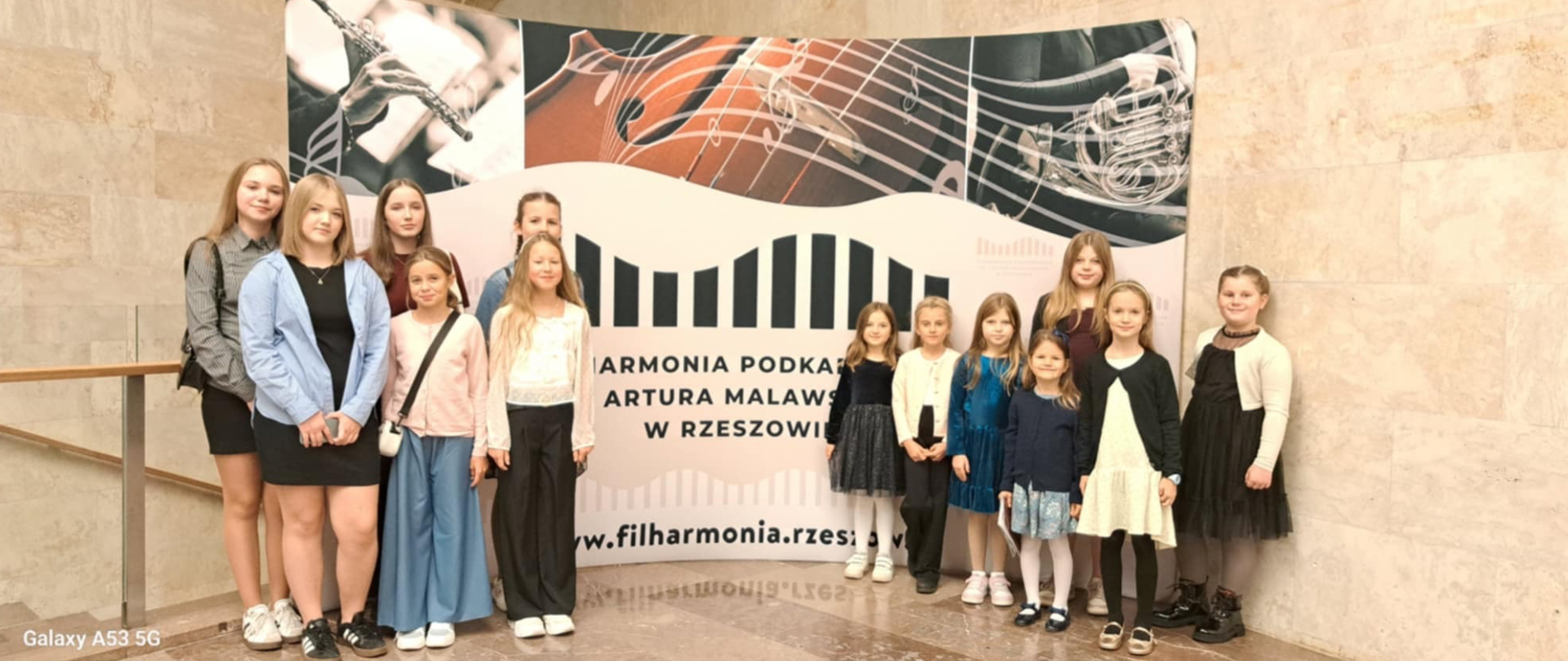 Zdjęcie grupowe przedstawiające jedenaście uczennic klasy fletu, stojących przed ścianką reklamową Filharmonii Podkarpackiej im. Artura Malawskiego w Rzeszowie. Dziewczęta są w różnym wieku, ubrane w codzienne stroje. W tle znajduje się baner promocyjny z grafikami instrumentów muzycznych, nut i stylizowanym logo Filharmonii z napisem: "FILHARMONIA PODKARPACKA IM. ARTURA MALAWSKIEGO W RZESZOWIE" oraz adresem strony internetowej.