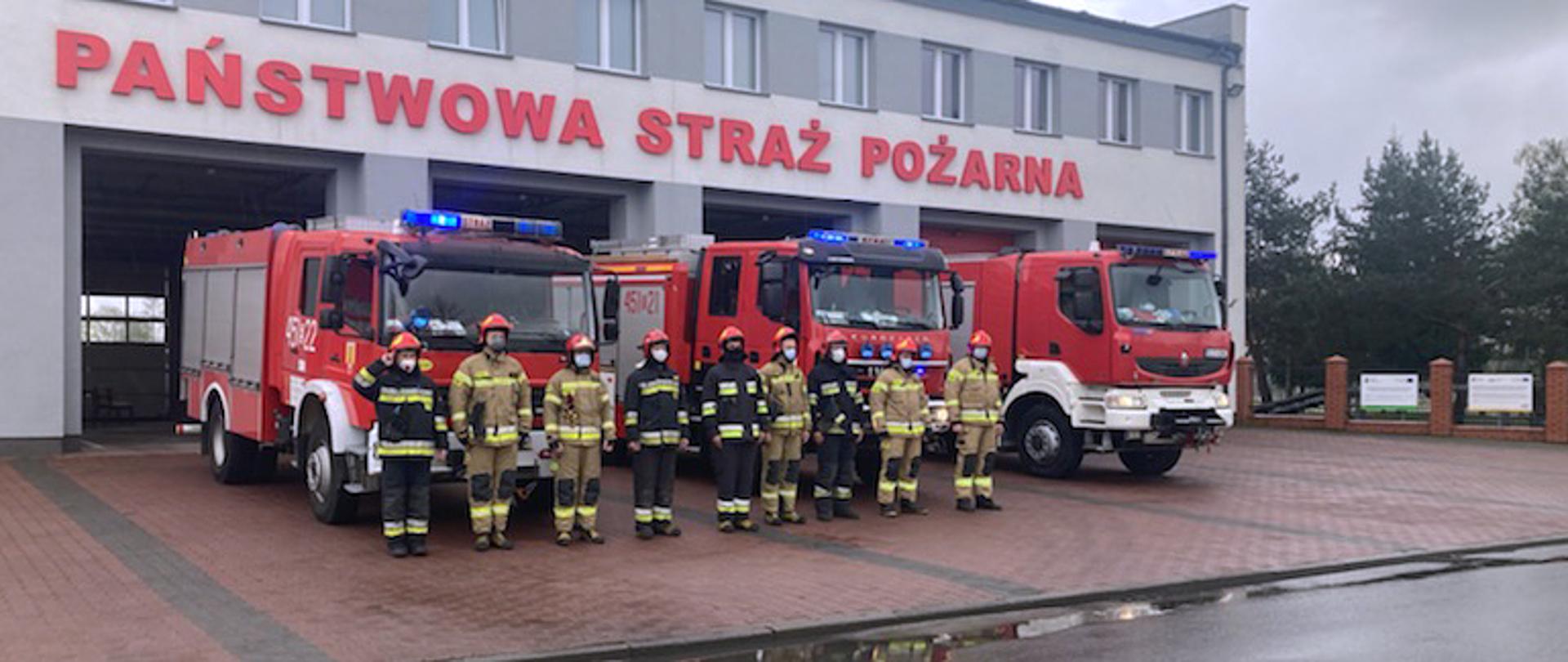 Zdjęcie przedstawia dziewięciu strażaków stojących w pozycji zasadniczej przed budynkiem Komendy Powiatowej PSP w Łowiczu. Jeden strażak po lewej stronie zdjęcia salutuje, za nimi stoją trzy pojazdy straży pożarnej z włączonymi sygnałami błyskowymi, wszyscy są ubrani w ubrania bojowe typu Moratex z odblaskami barwy szarej i żółtej, na głowie mają hełm a na twarzy maseczkę chirurgiczną. W tle budynek komendy oraz napis na froncie PAŃSTWOWA STRAŻ POŻARNA.