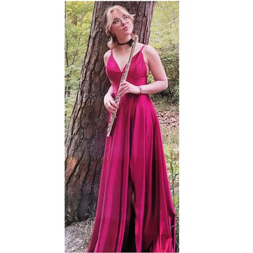 Zdjęcie kolorowe. Dziewczyna w czerwonej sukni, trzyma flet. W tle pień drzewa.