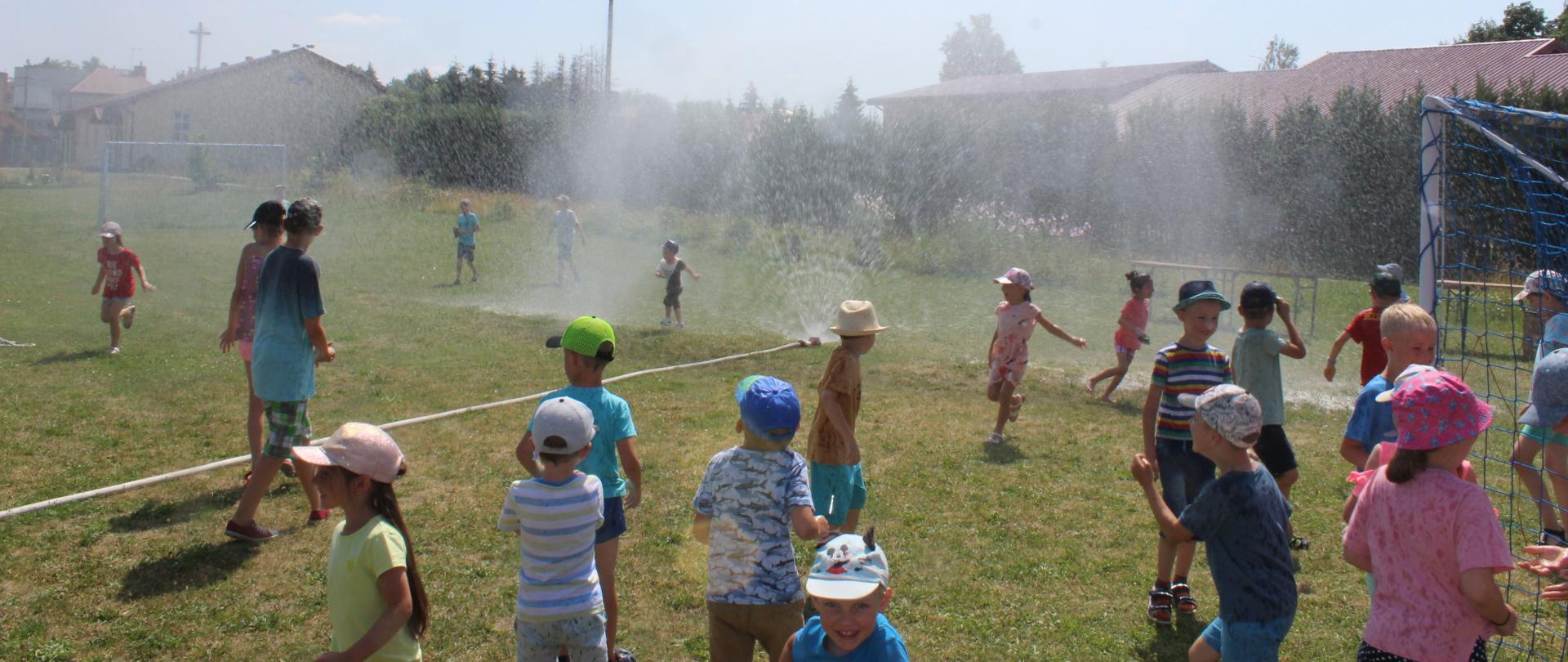 na zdjęciu widać dzieci w letnich strojach podczas zabawy przy kurtynie wodnej stworzonej przez strazaków