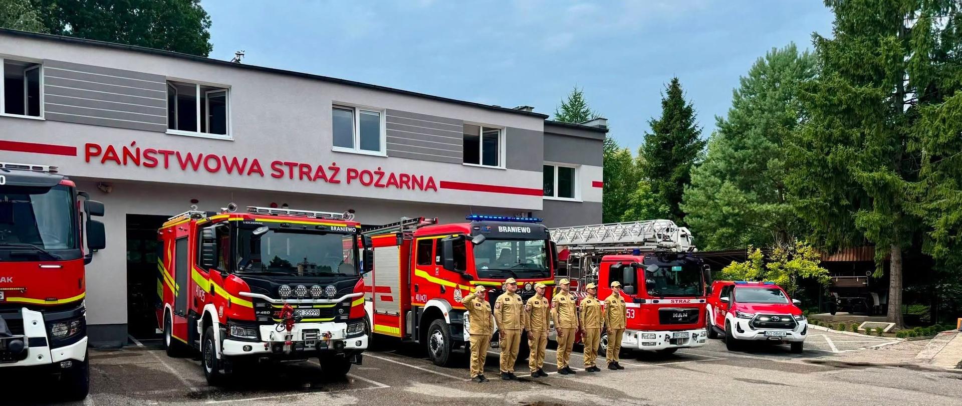 Przed budynkiem Komendy Powiatowej Państwowej Straży Pożarnej w Braniewie stoi grupa strażaków w piaskowych mundurach. Za nimi ustawiono kilka czerwonych samochodów ratowniczo-gaśniczych, w tym wóz z drabiną oraz samochód terenowy. W tle widać jasną elewację strażnicy z dużym czerwonym napisem „Państwowa Straż Pożarna” oraz wysokie zielone drzewa.