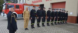Dzień flagi RP. Na tle wozu pożarniczego stoją strażacy ze zmian słuzbowych nr 2 i 3 JRG. Występuja w ubraniach koszarowych, a zastępca dowódcy JRG - w słuzbowym koloru piaskowego. Frontem do budynku stoi z kolei zastępca komendanta powiatowego PSP w mundurze wyjściowym.