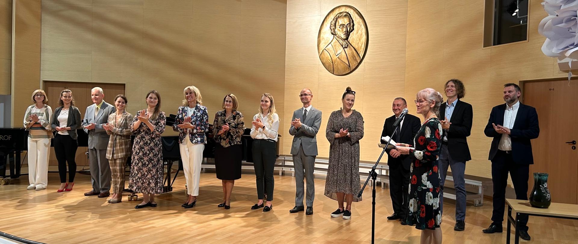 na scenie sali koncertowej stoją nauczyciele szkoły, klaszczą witając nowy rok szklony 