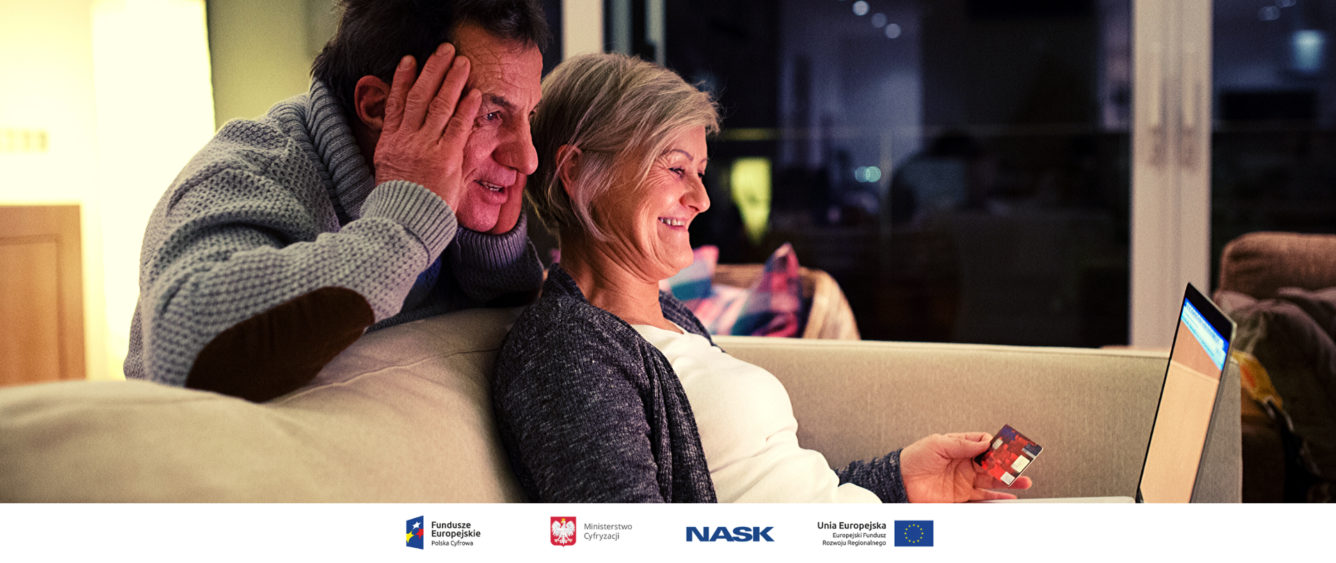 e-senior - program Ministerstwa Cyfryzacji Na kanapie siedzi starsza kobieta z laptopem na kolanach, w lewej ręce trzyma kartę płatniczą. Z tyłu o oparcie kanapy opiera się łokciami, podpierając głowę, starszy mężczyzna i również patrzy w ekran.