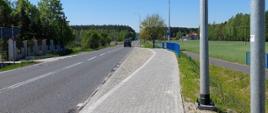 Chodnik z kostki brukowej wzdłuż drogi, w pobliżu zatoki drogowej i nowej infrastruktury oświetleniowej. Obok znajdują się pola, drzewa oraz pojedyncze zabudowania, a po jezdni poruszają się pojazdy.