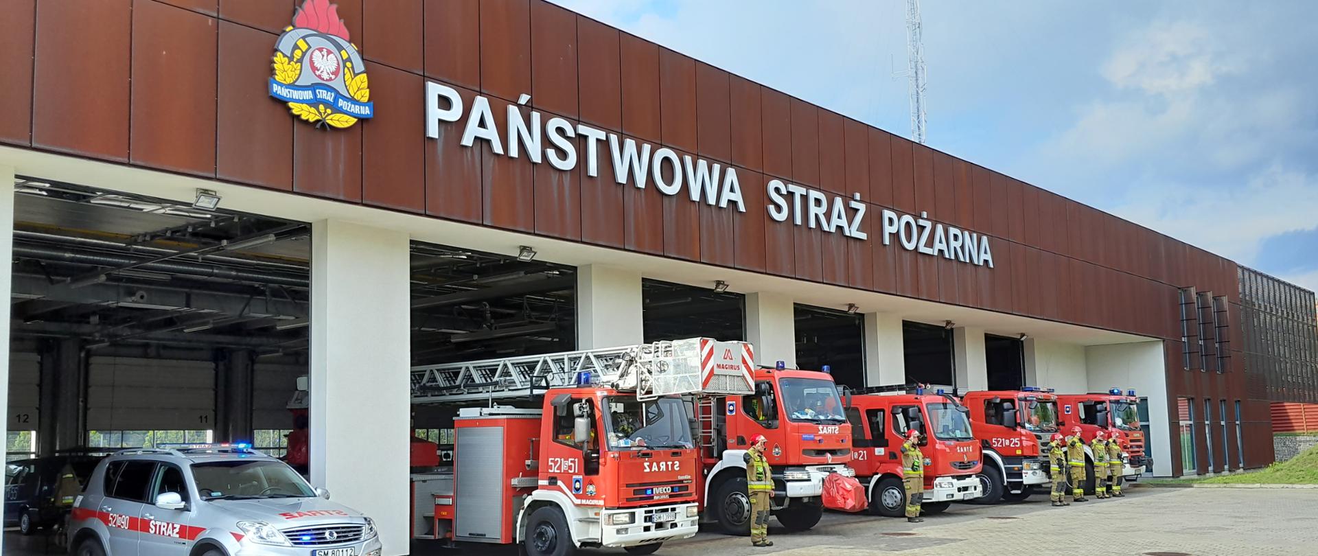 Samochody strażackie w bramie Komendy Miejskiej Państwowej Straży Pożarnej w Mysłowicach oraz strażacy stojący w nomexach przy samochodach bojowych.