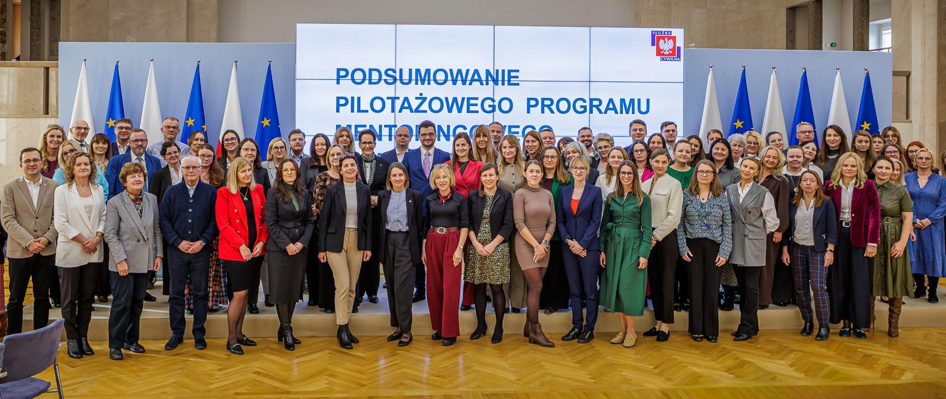 Bardzo duża grupa osób pozująca do zdjęcia, za nimi ekran z prezentacją oraz flagi PL i UE.