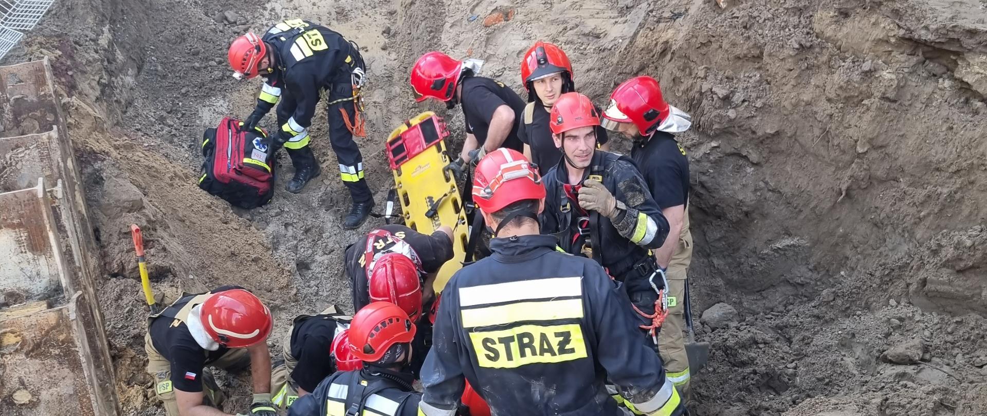 Strażacy stojący w wykopie przygotowują się do uwolnienia mężczyzny przysypanego ziemią