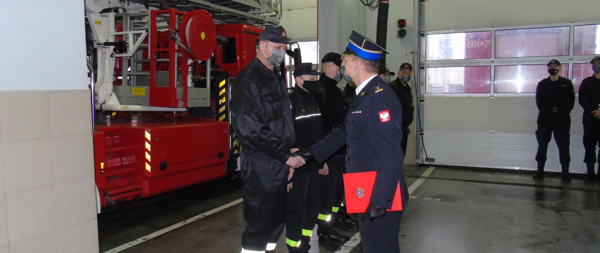 Uroczysta zbiórka w związku z odejściem na zaopatrzenie emerytalne asp. Marka Wawrzyniaka. Na pierwszym planie Komendant Powiatowy wręcza strażakowi decyzję o przejściu na zaopatrzenie emerytalne. Na drugim planie samochód specjalny auto drabina. 