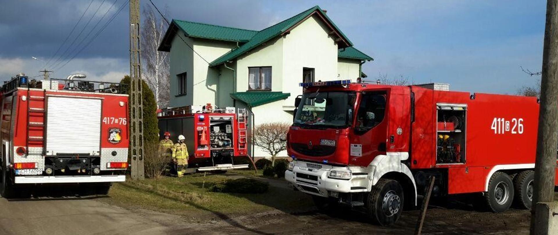 Przed zielonym budynkiem mieszkalnym na drodze gruntowej stoją samochody strażackie uczestniczące w działaniach. 