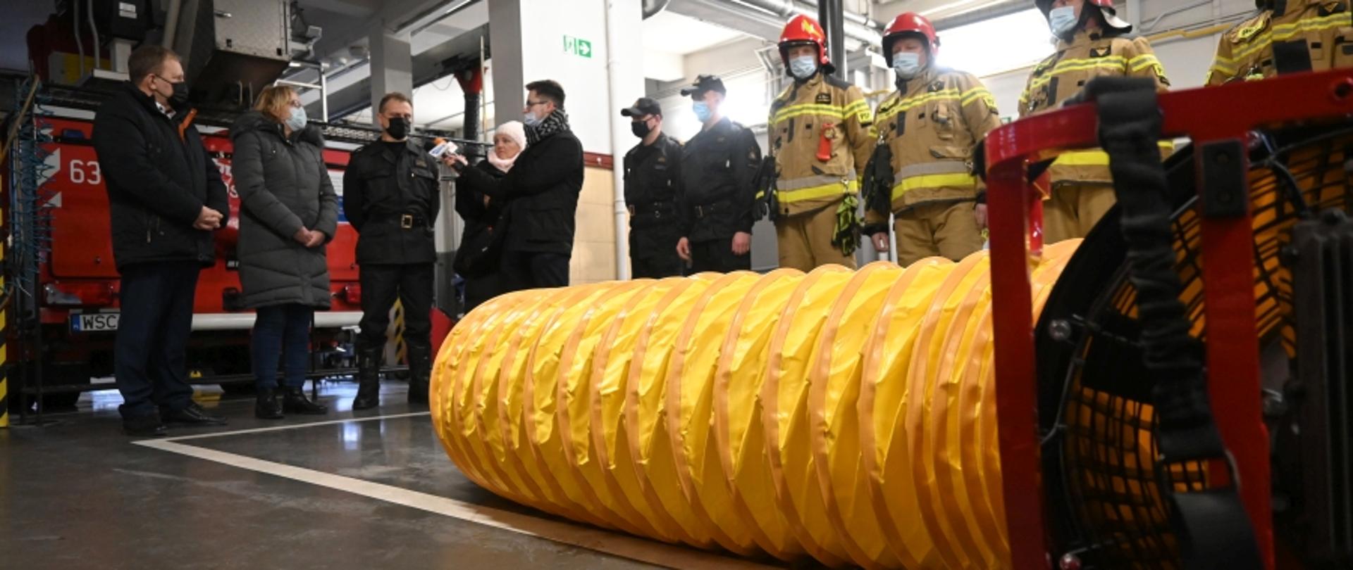 Na pierwszym planie wentylator z zamocowanym do niego żółtym, karbowanym rękawem. Na drugim planie komendant powiatowy w obecności wicestarostów wypowiada się do mikrofonów lokalnych dziennikarzy. W tle strażacy stojący w szeregu.