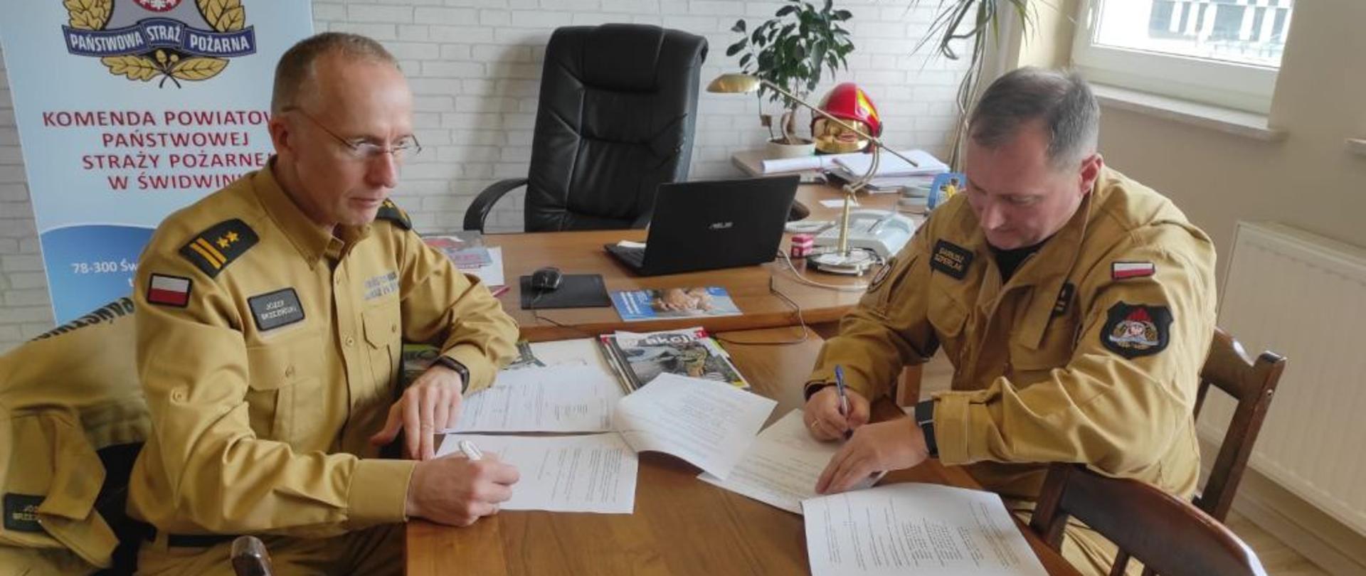 Na zdjęciu dwaj strażacy w piaskowym umundurowaniu. Strażacy podpisują dokumenty operacyjne. Jeden ze strażaków starszy w okularach drugi młodszy.