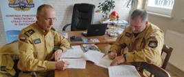 Na zdjęciu dwaj strażacy w piaskowym umundurowaniu. Strażacy podpisują dokumenty operacyjne. Jeden ze strażaków starszy w okularach drugi młodszy.