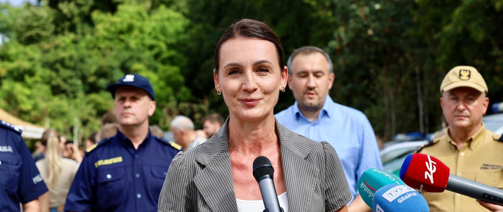 Kobieta (wojewoda pomorska) przemawia podczas konferencji prasowej na Pikniku Rekrutacyjnym Służb Mundurowych. Za nią stoją mężczyźni w mundurach. W tle widoczne są drzewa. 